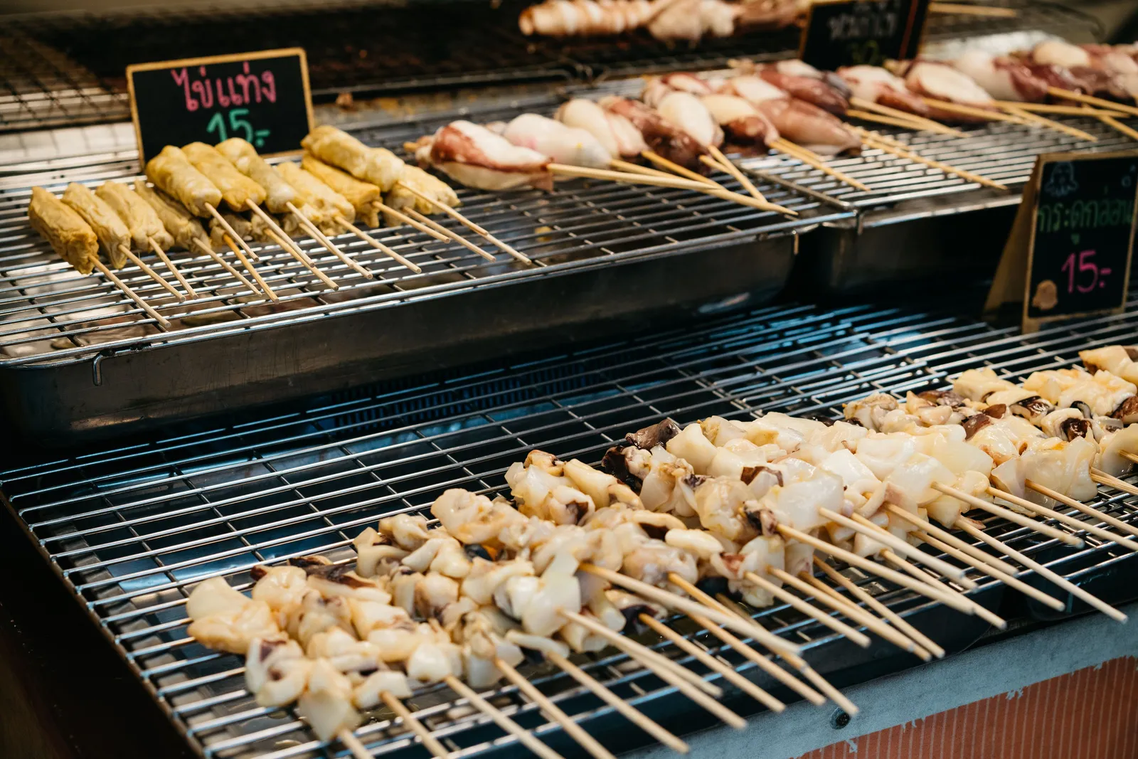 Grilled seafood skewers on a charcoal grill at a market stall with a Thai price sign, Amphawa, Thailand