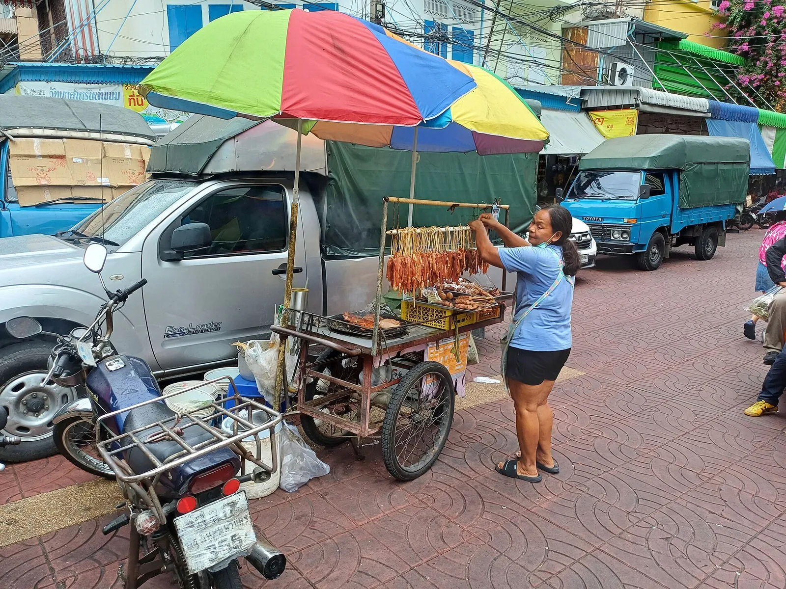 A street vendor grilling skewers at a cart under a bright umbrella on a Bangkok street, Thailand
