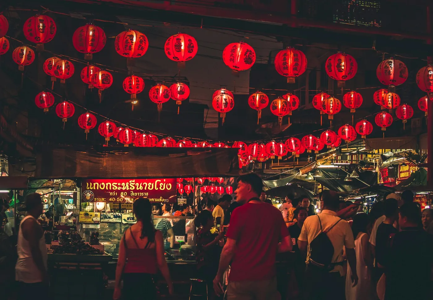 A crowded night market street in Bangkok hung with red lanterns and neon signs, Thailand