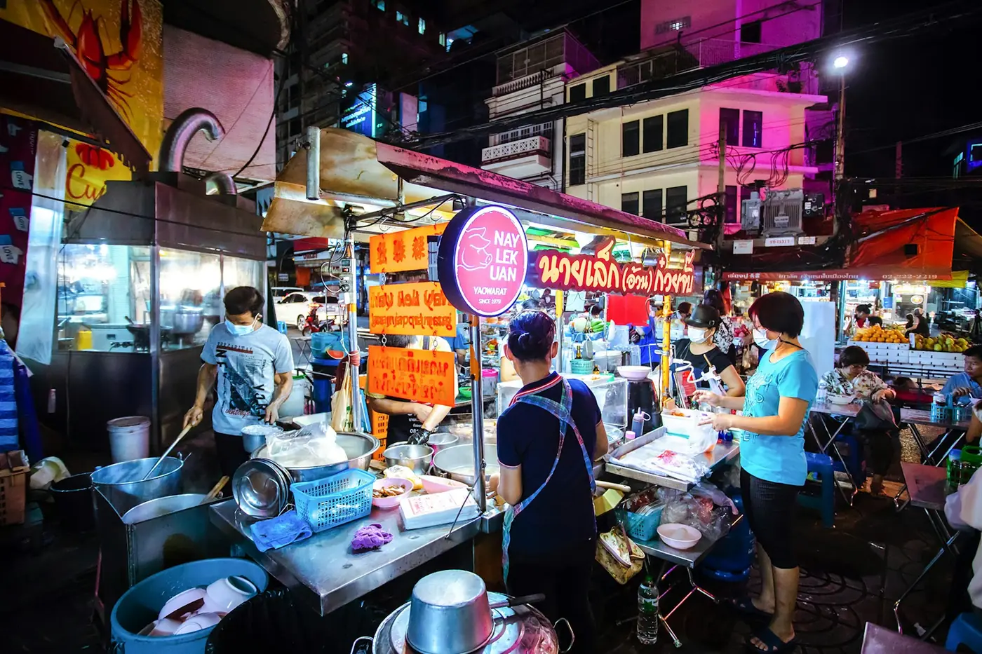 A busy noodle stall in Bangkok's Chinatown in daylight, with customers waiting at the counter for rolled noodle soup