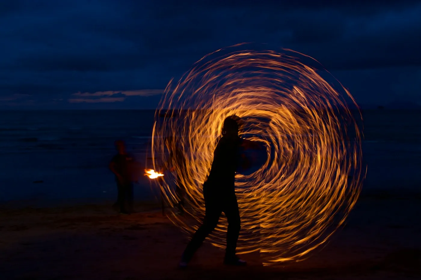 A fire performer drawing a ring of flame beside a burning full moon party sign on a beach in Thailand at night