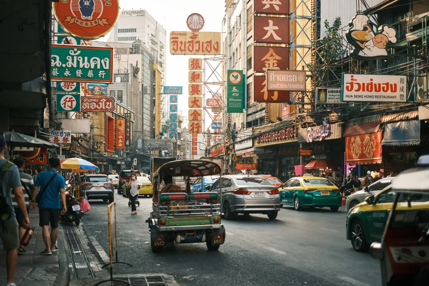 Khao San Road in Bangkok by day, lined with bar signs and market stalls, tuk-tuks parked along the street under a clear sky