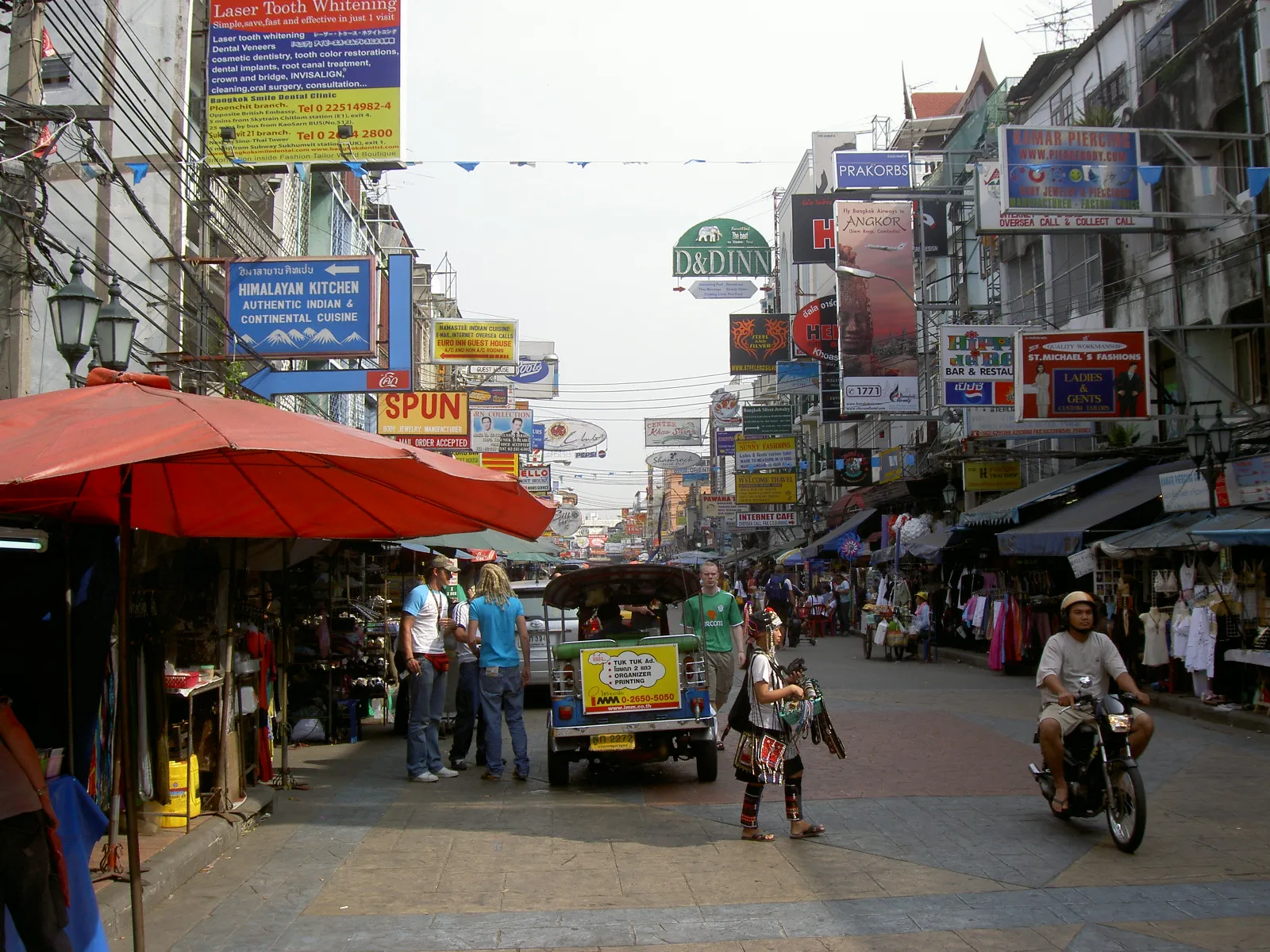 Khao San Road in Bangkok during the day, overhead shop and restaurant signs, a market umbrella and a tuk tuk on the street