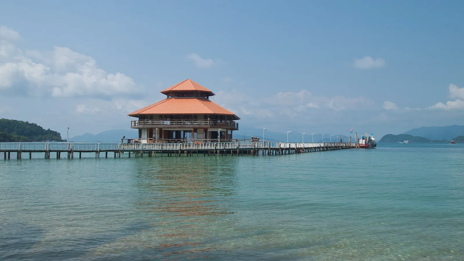 A wooden pier leading to a pavilion with a tiered orange roof over turquoise water, forested hills across the bay on Koh Chang, Thailand