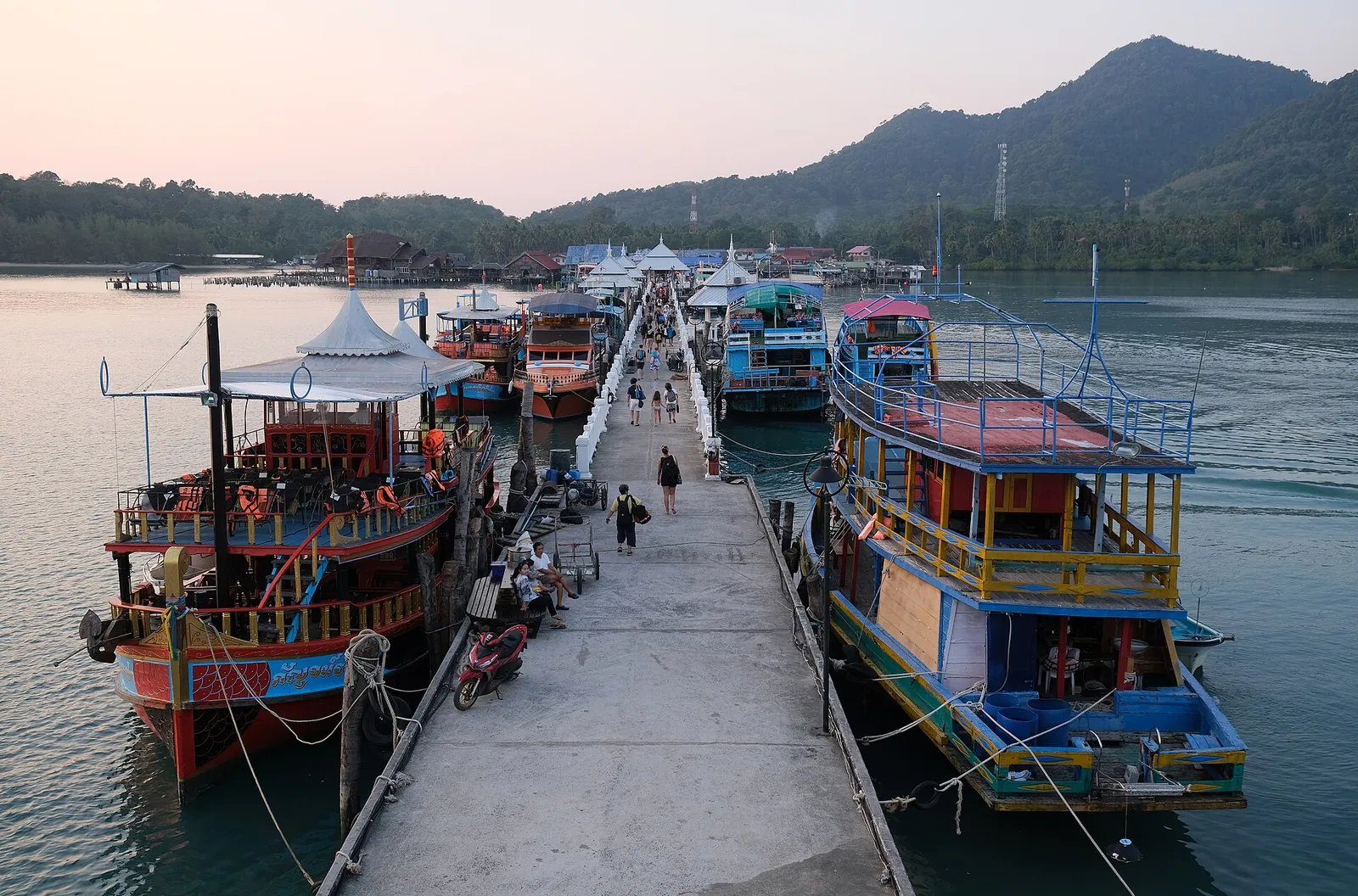 Traditional wooden tour boats moored along a pier as visitors walk past at dusk, forested hills behind on Koh Chang, Thailand