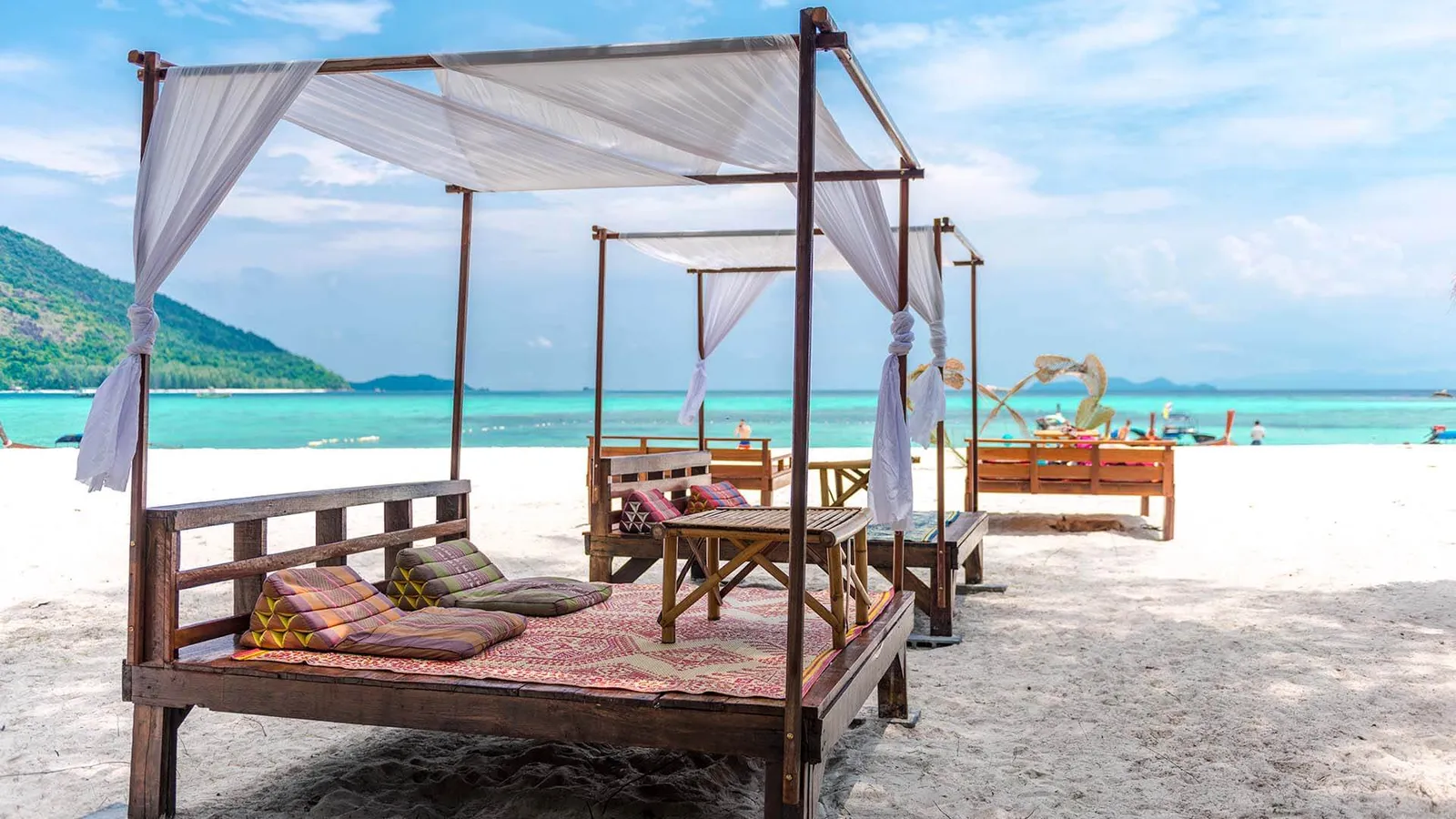 Canopied wooden day beds with cushions on a white sand beach in Koh Lipe, Thailand, turquoise water and a green hillside behind