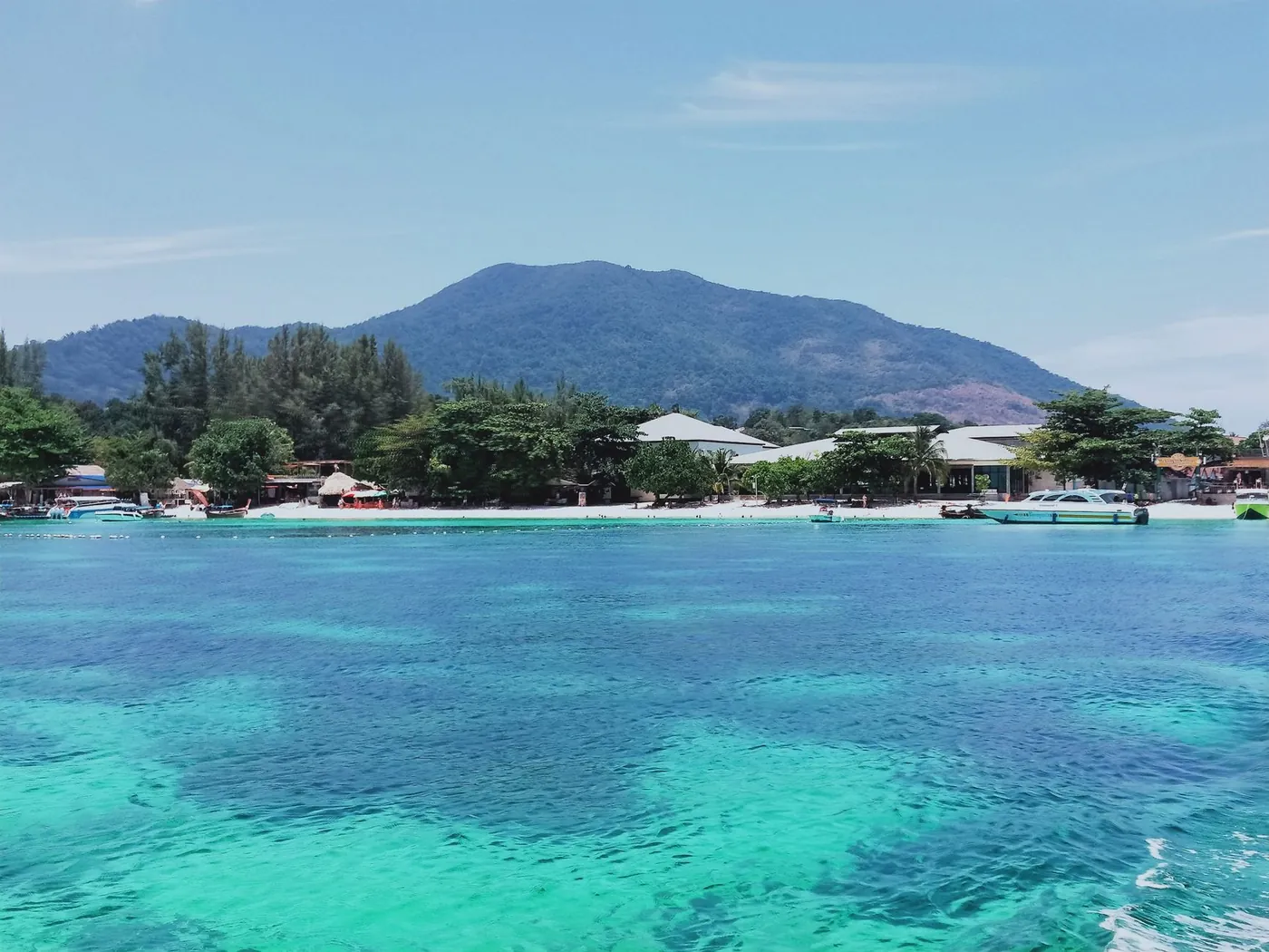 View across turquoise water toward Koh Lipe's forested hills and a cluster of waterfront buildings, with boats moored close to shore