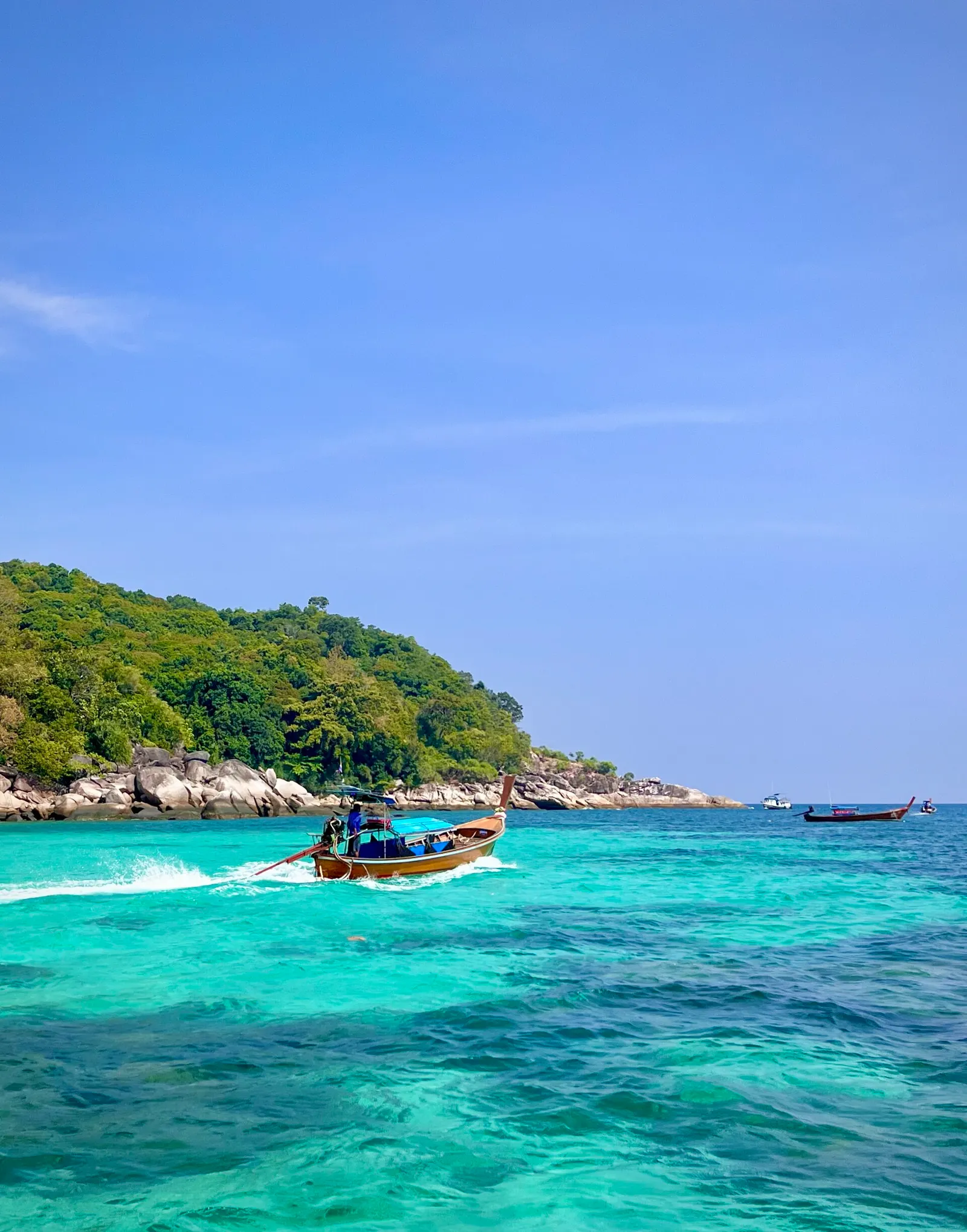 A wooden longtail boat crossing turquoise water near a forested, rocky headland in the Adang-Rawi Archipelago, Thailand
