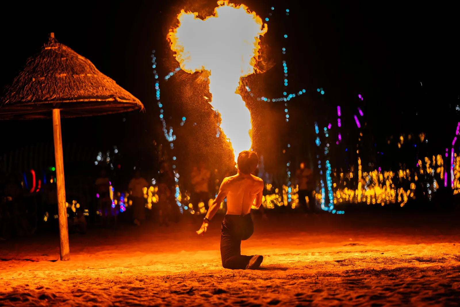A fire performer kneeling on the sand at a night beach party in southern Thailand, breathing a tall column of flame beside a thatched beach umbrella and strings of coloured lights