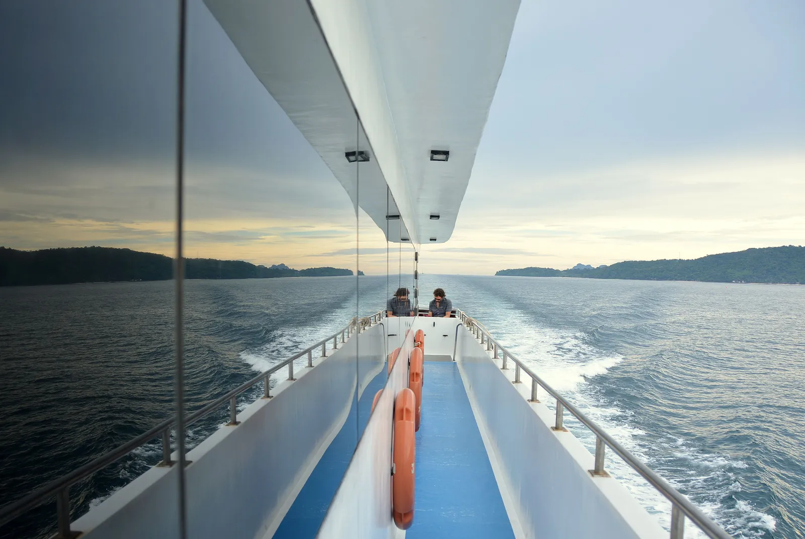 The stern deck of a ferry in the Gulf of Thailand with two passengers seated at the back, orange life rings mounted on the rail, and hills visible on both sides of the water