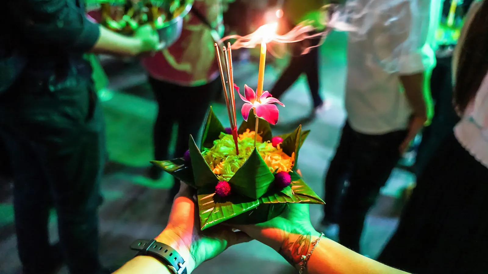 Hands holding a banana leaf krathong with a lit candle, incense sticks and an orchid at night in Thailand