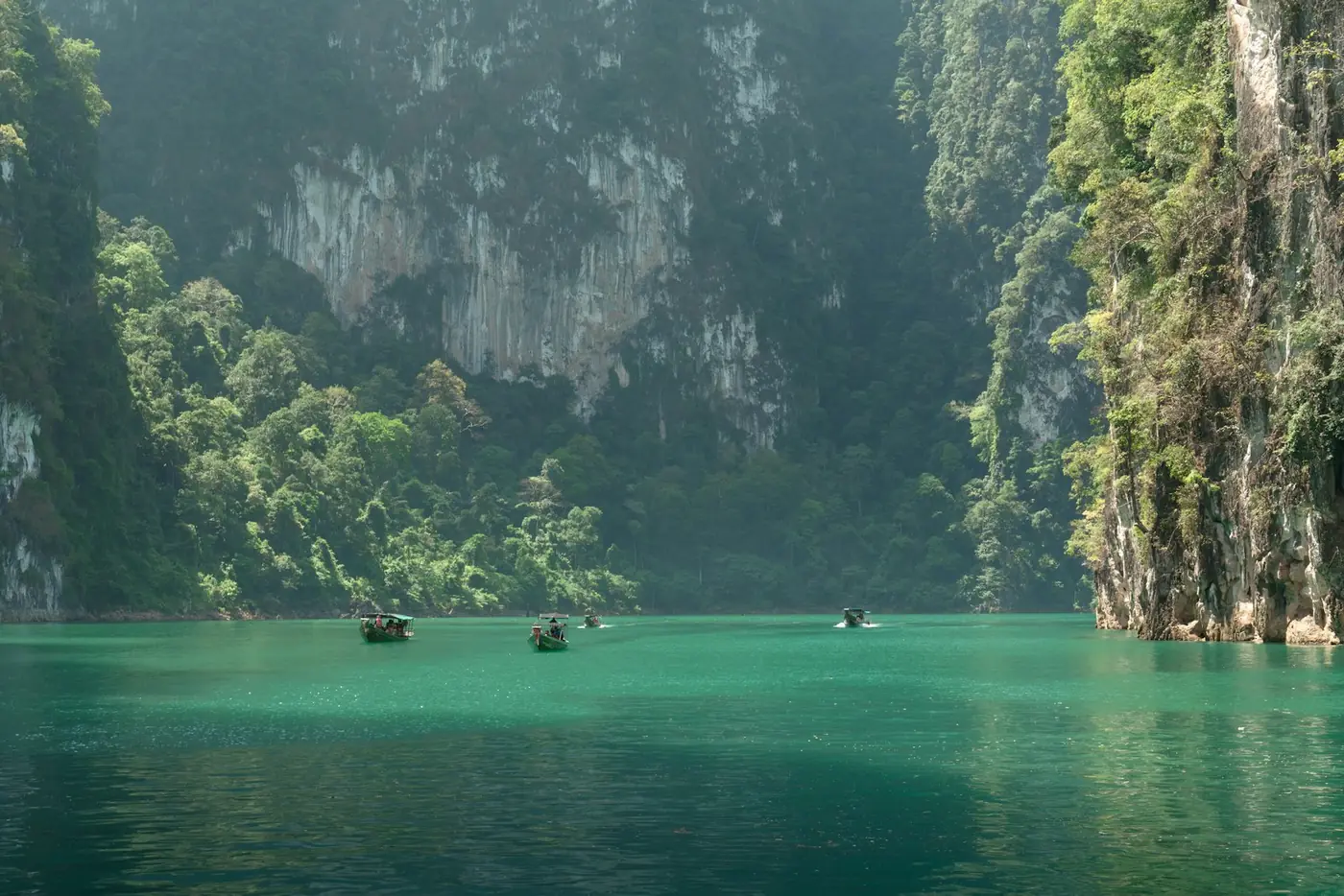 Small boats beneath towering limestone cliffs on calm water, a landscape common across Thailand's karst regions