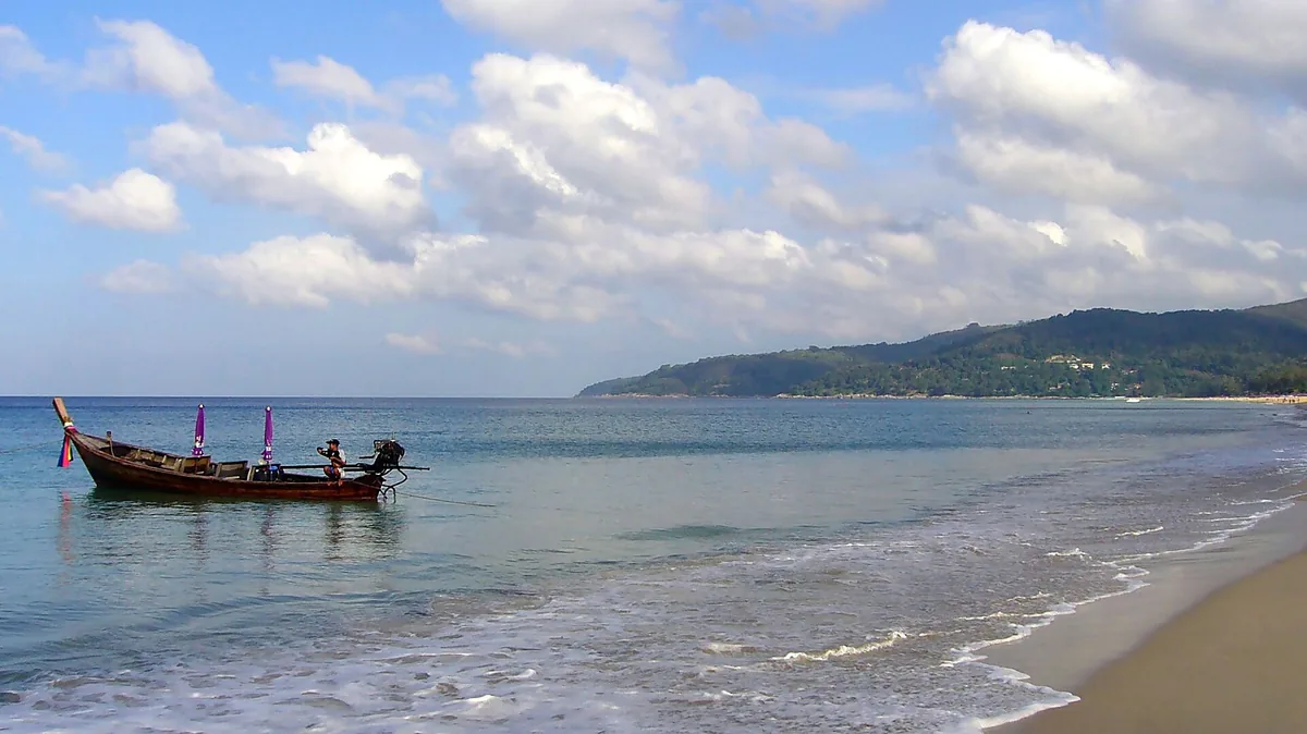 A longtail boat moored in shallow water beside a wide sandy beach, with a green headland closing the bay