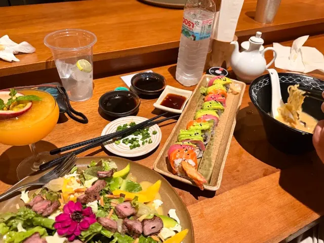 A colorful sushi roll, a beef salad and a bowl of ramen on a wooden counter