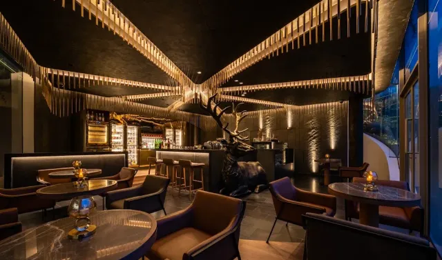 A dark lounge with a lit slatted wooden ceiling, a large deer sculpture and round armchairs