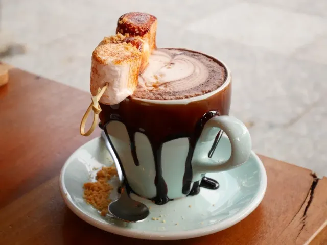 A cup of hot chocolate with a toasted marshmallow skewer and chocolate dripping onto the saucer