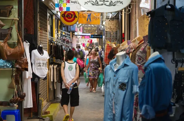 A narrow market lane lined with colourful clothing stalls and shoppers walking between them