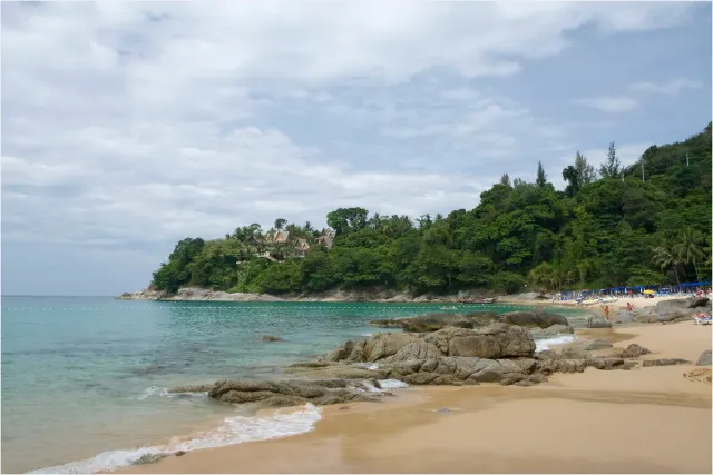 Rocks on golden sand facing a green headland with houses, pale sea on the left