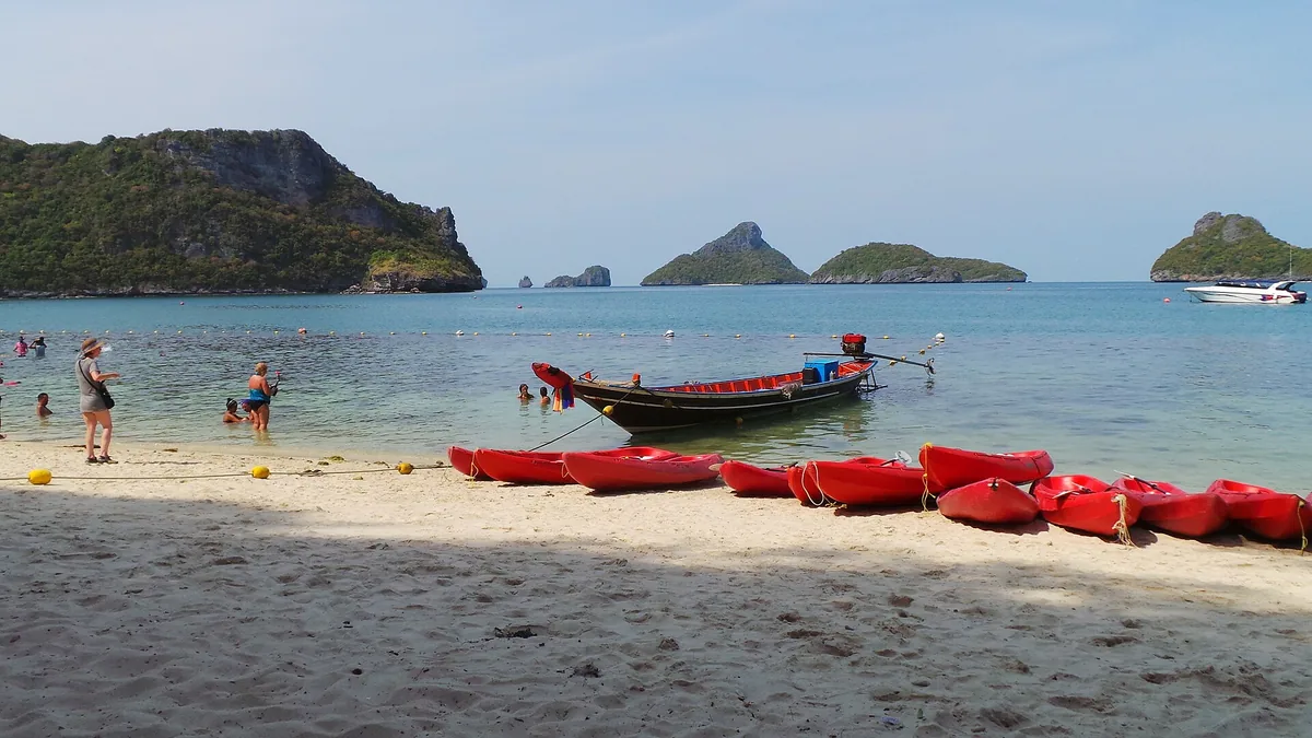 Red kayaks and a longtail boat on a white sand beach, facing green limestone islands in shallow water