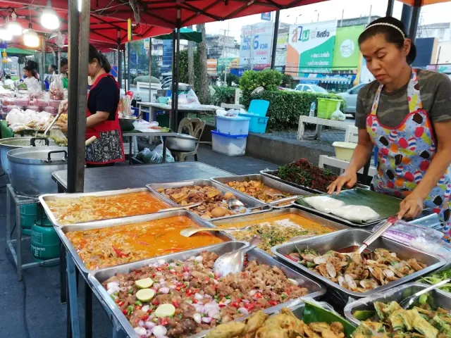 A ready food stall with metal trays full of cooked dishes, a vendor serving a portion