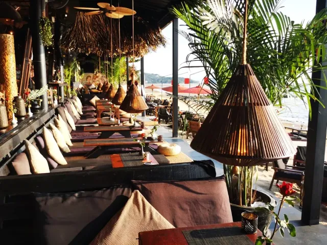 A beach lounge with low mattresses, cushions and rattan shades above, and the sea in the last light