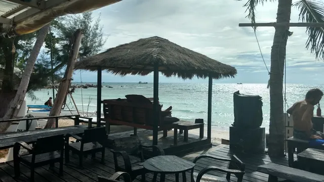 A thatched gazebo with a wooden bench on a deck facing the beach and the calm sea