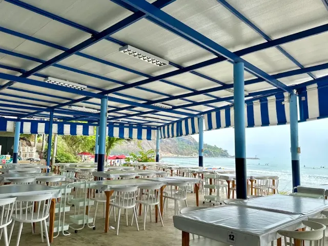 A hall roofed in blue and white stripes with white tables and an open beach to the side