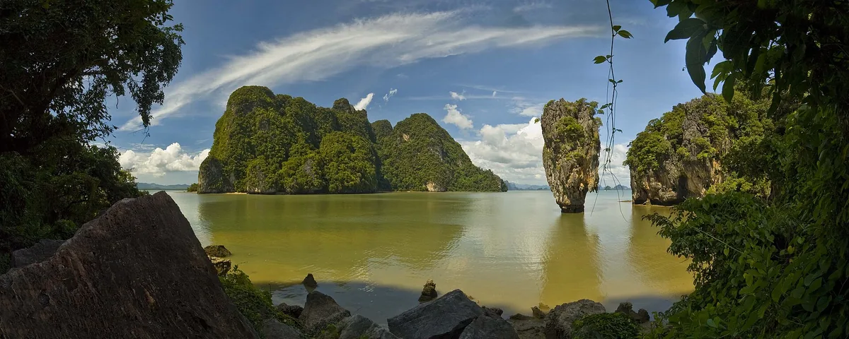 Forested limestone cliffs rising from calm green water with a narrow needle rock to the side