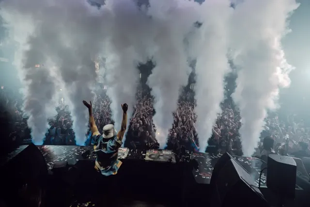 A DJ raising his arms before a dense crowd, white smoke jets behind him
