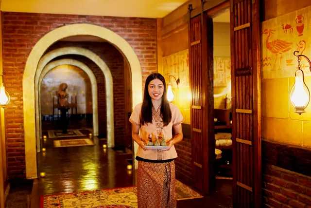 A therapist in traditional dress serves a tray in a brick archway corridor lit by lanterns
