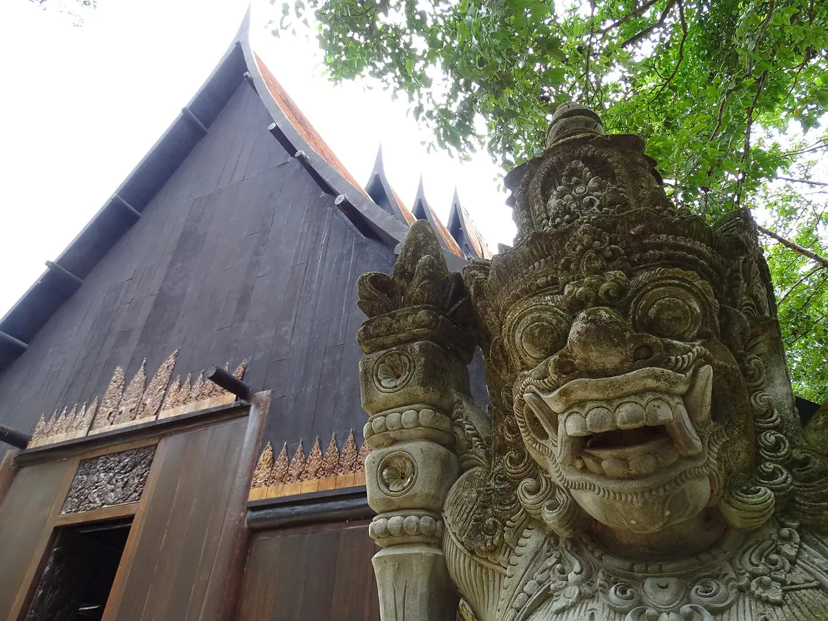 A dark timber building with a steep pitched roof and a carved guardian figure in front, among green trees