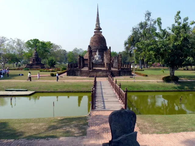 A white chedi on a small lake island with a stone causeway leading to it between trees