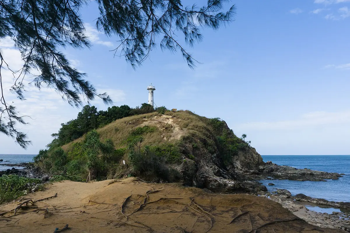 A rocky wooded cape with a small lighthouse on its summit, blue sea on both sides