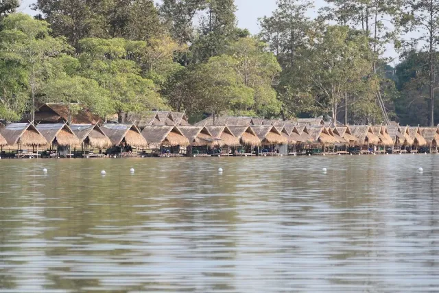 A row of thatched huts on a quiet lake shore, forest behind and gray water