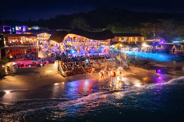 A beach club lit up at night, with a fire show on the sand by the waterline