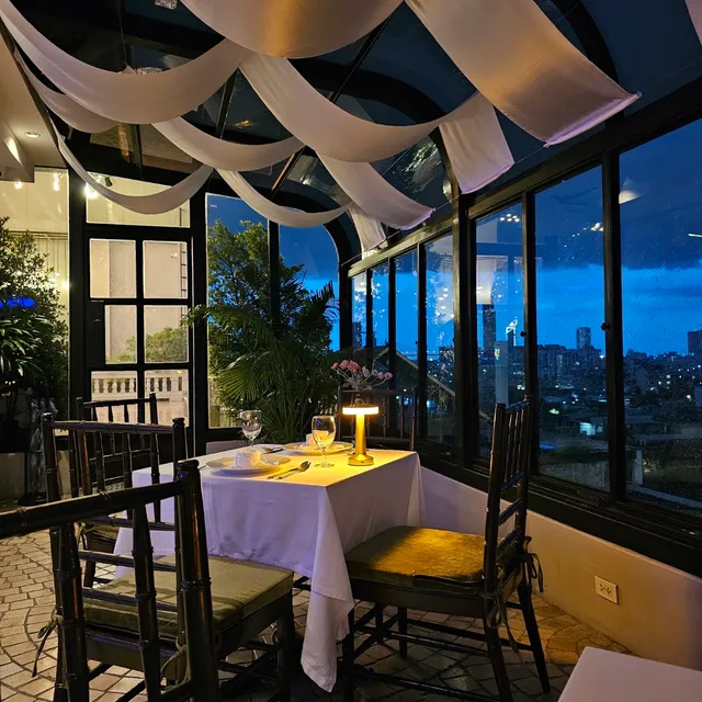 A table set with a white cloth in a glass conservatory, with city lights in the windows in the evening