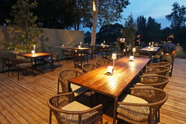 A wooden deck at dusk with long tables, woven chairs and table lamps