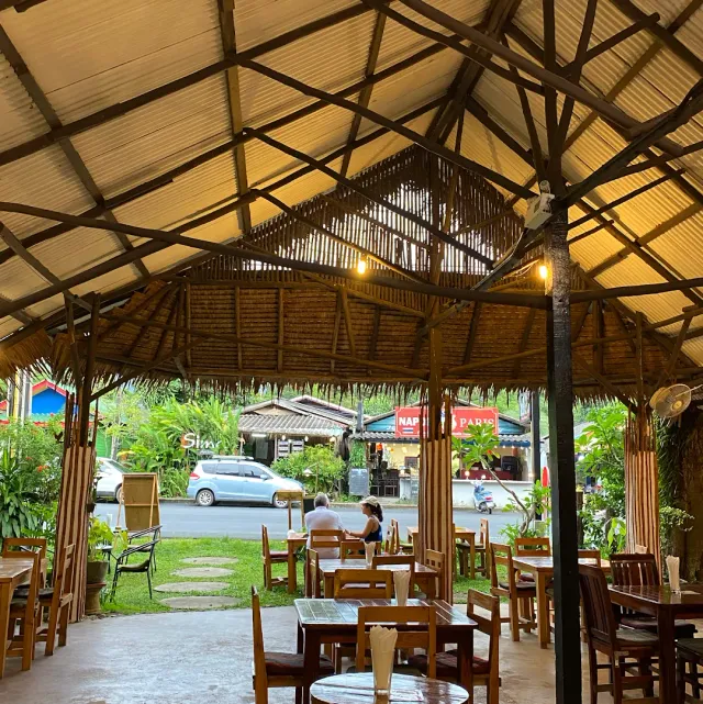 A pavilion roofed in tin and thatch with wooden tables and a view to the road
