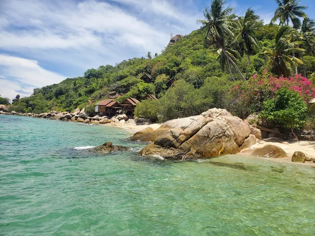Clear turquoise water and granite boulders before a small beach with huts on the hill