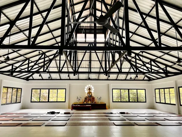 A large meditation hall with black steel beams, rows of mats and a golden statue