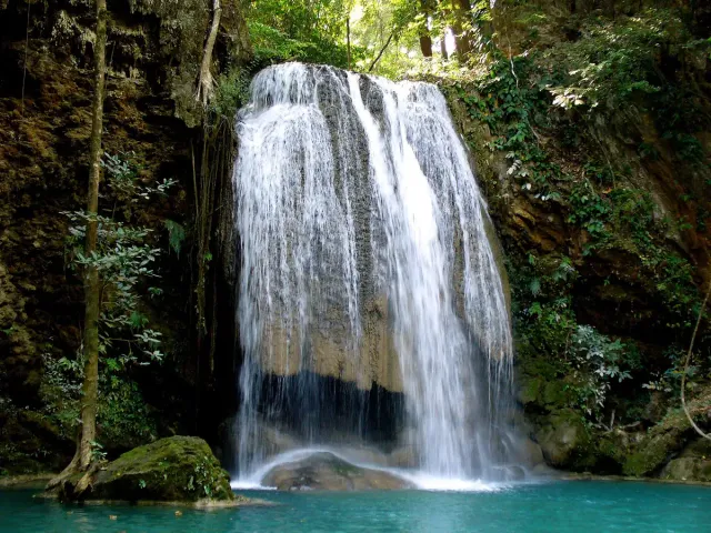 A tiered waterfall spilling over limestone rocks into a green pool inside dense forest