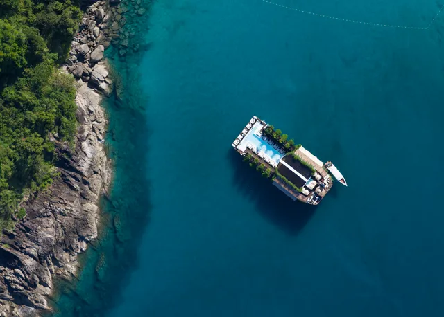 An overhead view of a floating platform with a pool in turquoise water beside a rocky shore