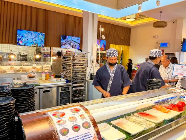 Chefs in patterned caps behind a sashimi counter with fish slices, and screens above them