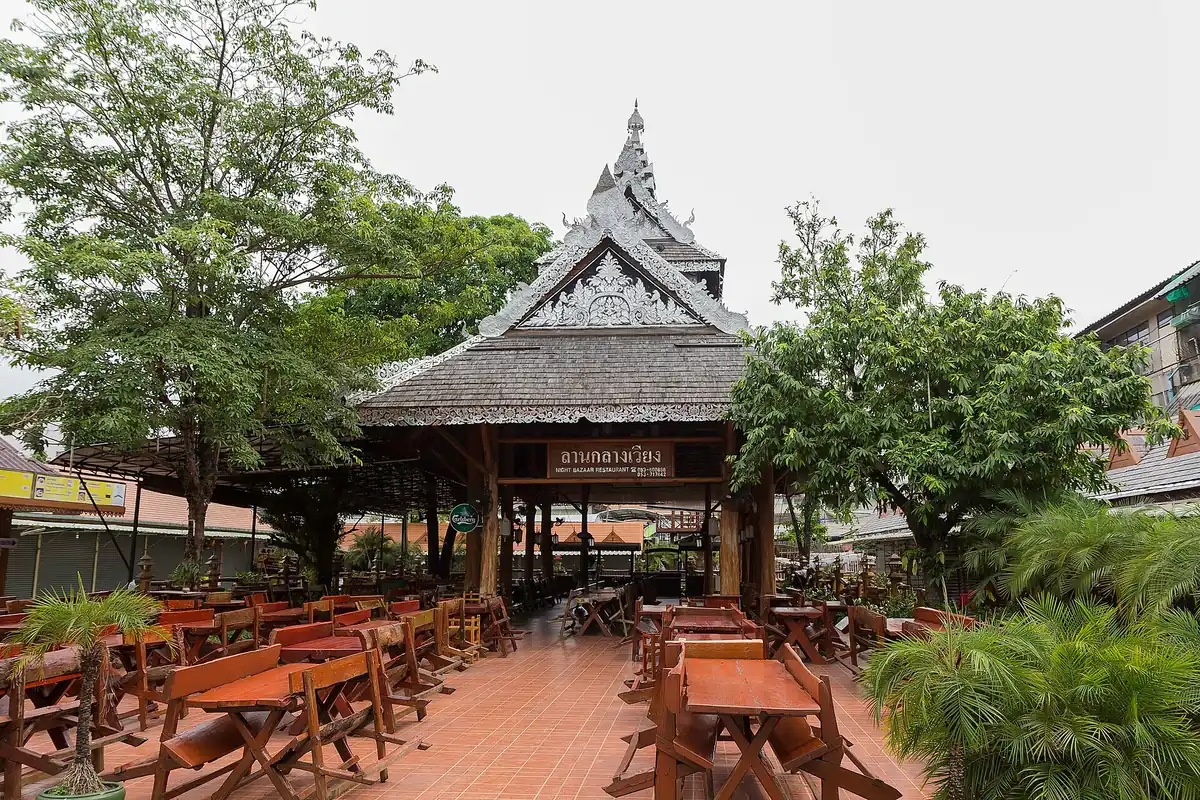 A Lanna style wooden pavilion with a carved pointed roof, and tables and benches on the terrace in front