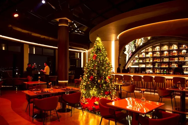 A decorated Christmas tree in the middle of a hall with red tables and a lit arched bar