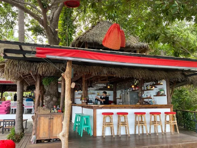 An outdoor bar under a thatched roof with tall red stools among large trees