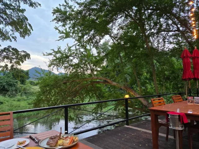 A wooden terrace above a stream with a served plate in front, red umbrellas and hills in the background