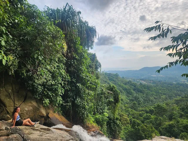 A hiker sits on a rock at the top of a waterfall, facing a green jungle valley to the horizon
