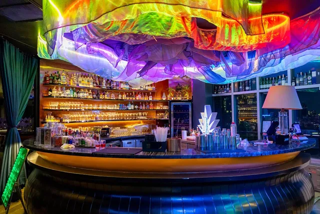 A round bar counter beneath a ceiling installation of glowing rainbow fabric