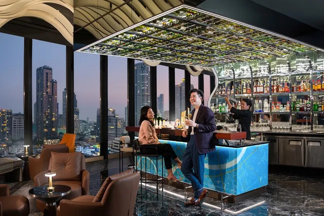A couple at a blue-lit bar counter, facing tall windows and city towers at dusk